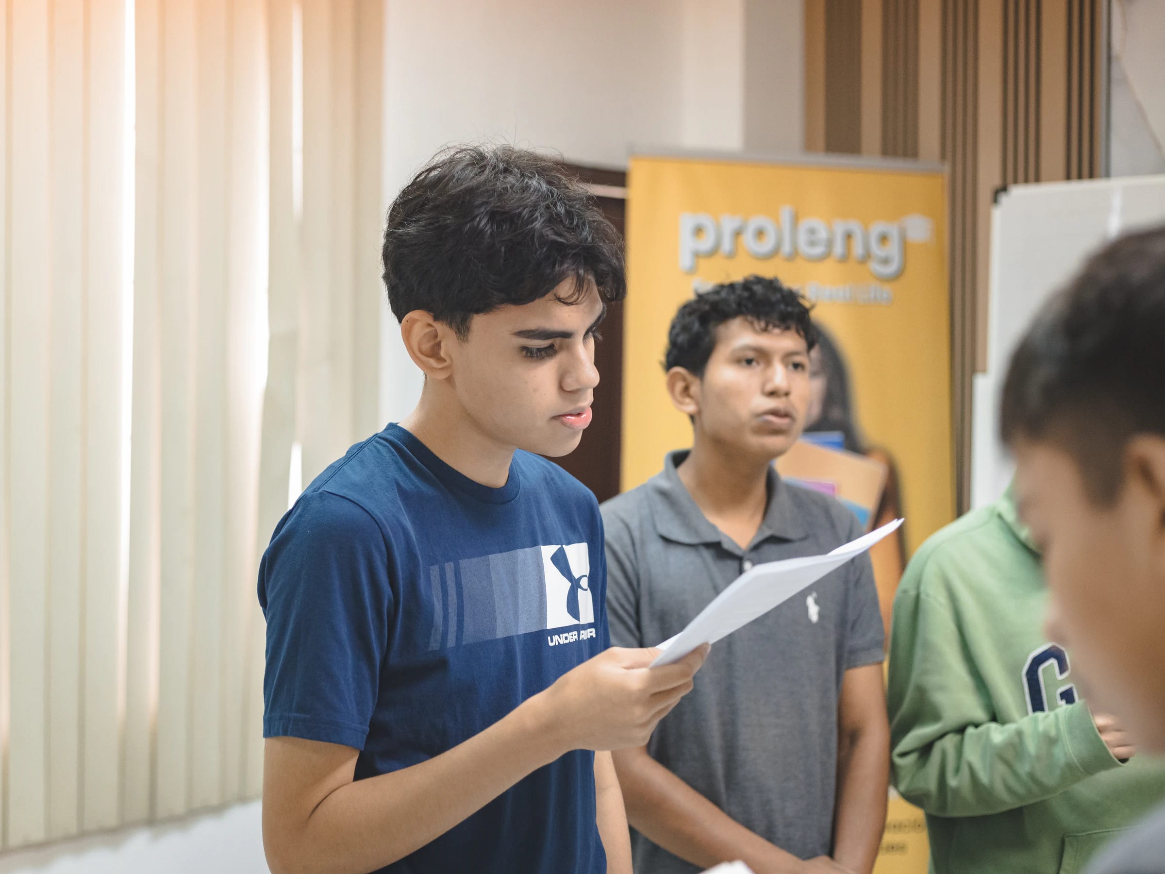 Estudiantes de ProLeng leen en voz alta con hojas en la mano durante una clase, con el banner de ProLeng al fondo