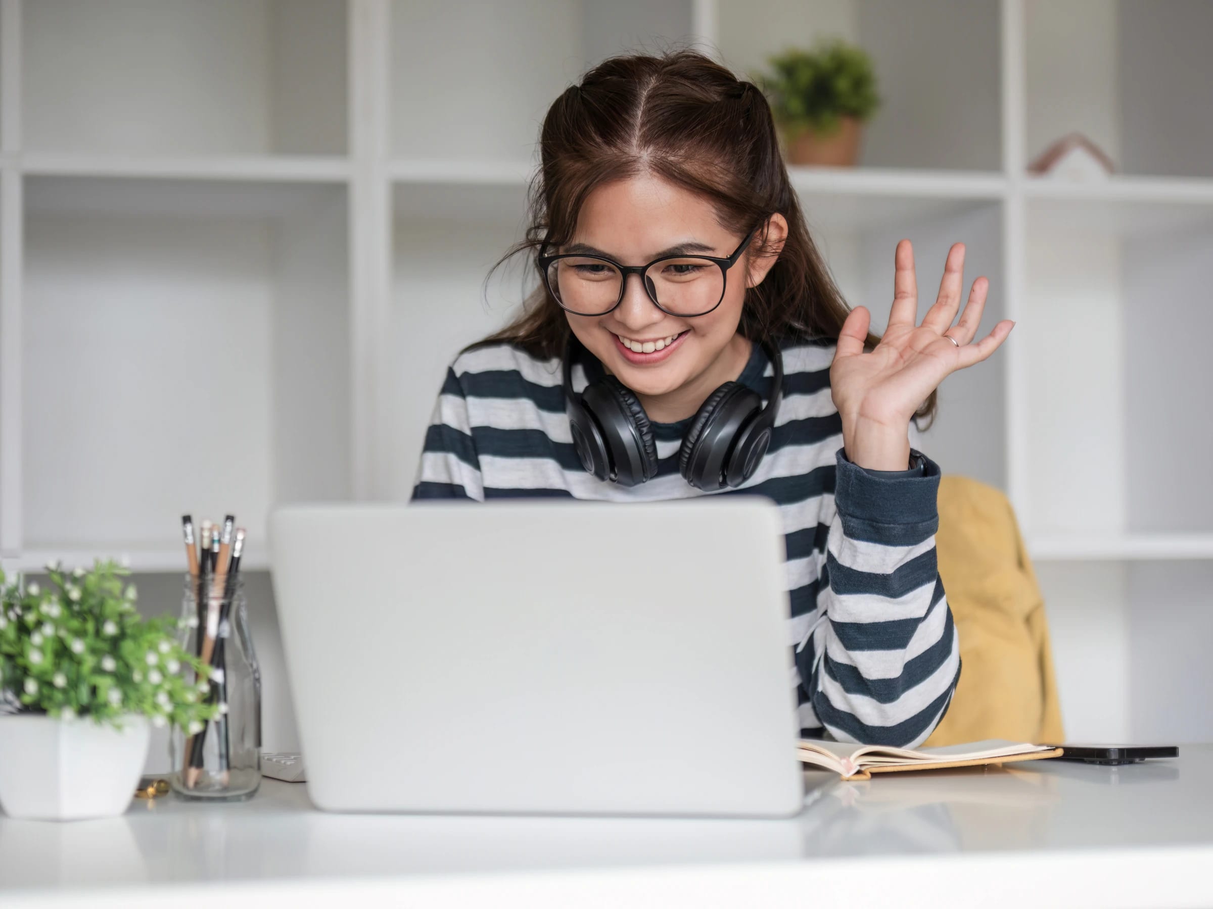 Mujer joven con lentes y audífonos al cuello saluda con la mano frente a su laptop durante una clase online